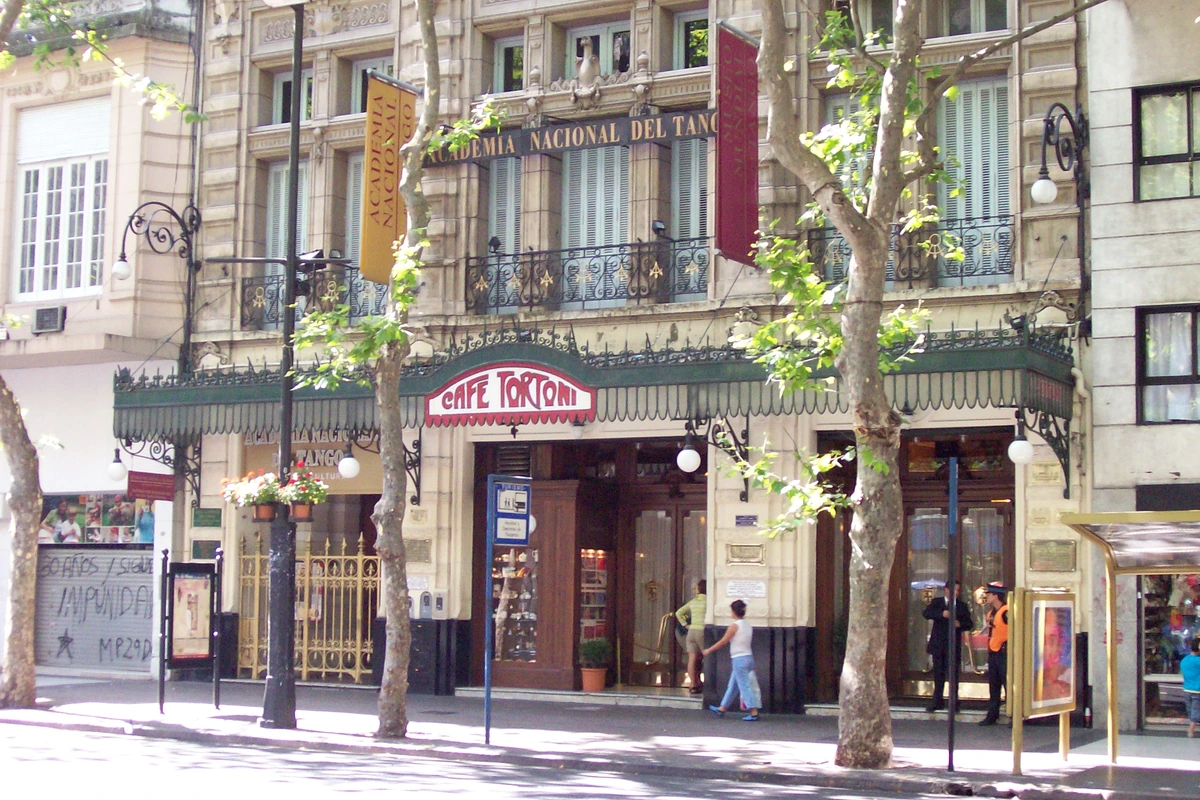 Six adults walk together on a sunlit path beside trees and low buildings