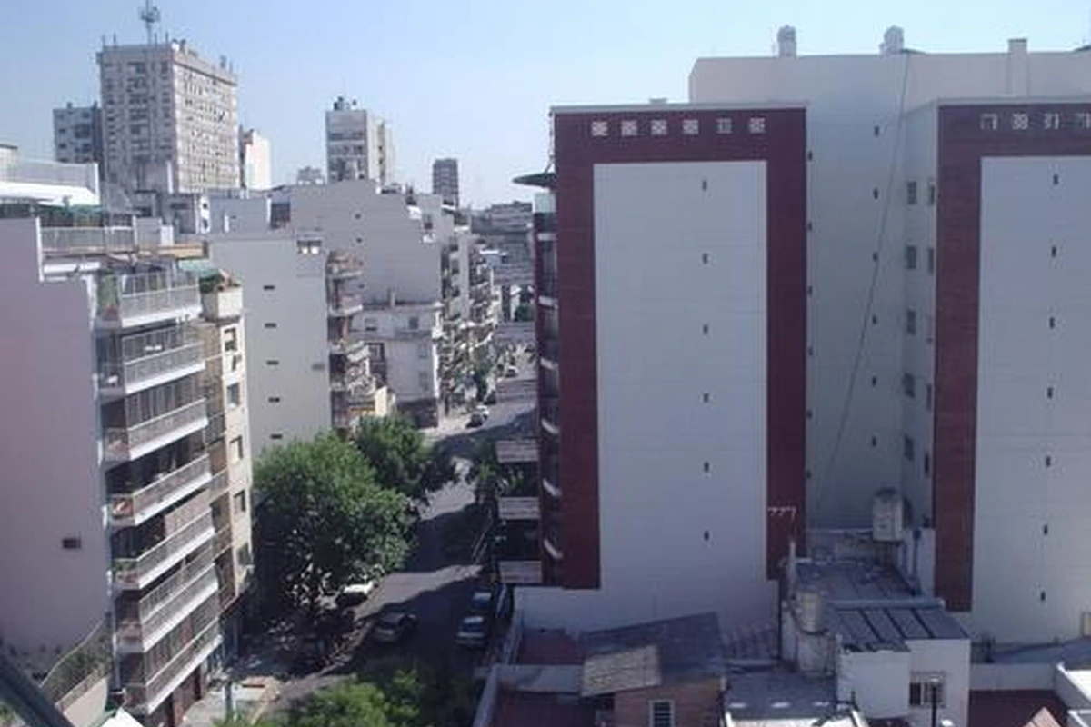 A sunny urban street has mid-rise apartment buildings, balconies, trees, parked cars, and pedestrians on the sidewalks